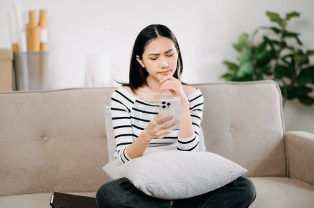 Téléchargez les photos : Jeune femme asiatique se détendre sur un canapé confortable à la maison SMS messagerie sur smartphone, fille souriante utiliser téléphone portable achats en ligne de la maison sur le canapé - en image libre de droit