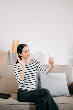 Téléchargez les photos : Jeune femme asiatique se détendre sur un canapé confortable à la maison SMS messagerie sur smartphone, fille souriante utiliser téléphone portable achats en ligne de la maison sur le canapé - en image libre de droit