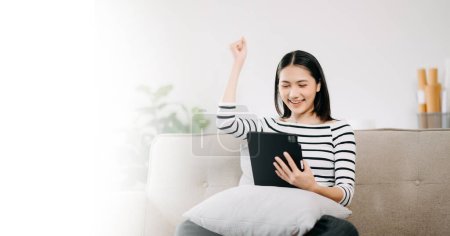 Foto de Joven mujer asiática relajarse en cómodo sofá en casa utilizando tableta digital, chica sonriente utilizar el teléfono celular de compras en línea desde la tableta de la computadora en el sofá - Imagen libre de derechos