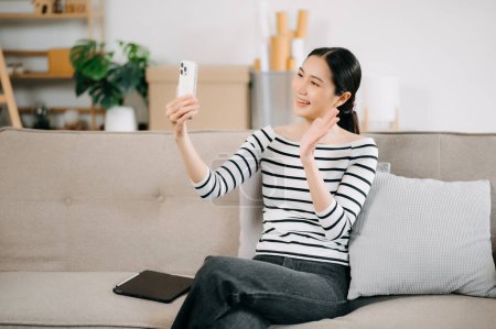 Foto de Feliz joven asiático mujer usando smartphone mientras sentado en sofá en casa - Imagen libre de derechos