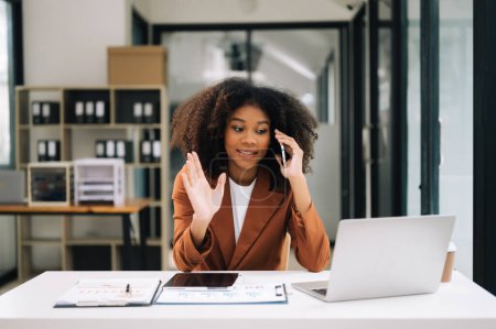 Téléchargez les photos : Femme africaine d'affaires Parler au téléphone et utiliser un ordinateur portable avec un sourire tout en étant assis au bureau moderne - en image libre de droit