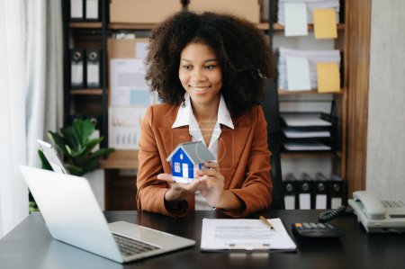 Téléchargez les photos : Jeune agent immobilier travailleur travaillant avec ordinateur portable à la table dans le bureau moderne et le modèle de petite maison - en image libre de droit