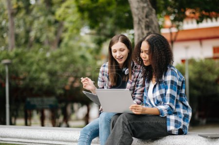 Téléchargez les photos : Jeunes étudiantes se concentrant sur le projet scolaire, utilisant un ordinateur portable, discutant et travaillant ensemble au parc du campus - en image libre de droit