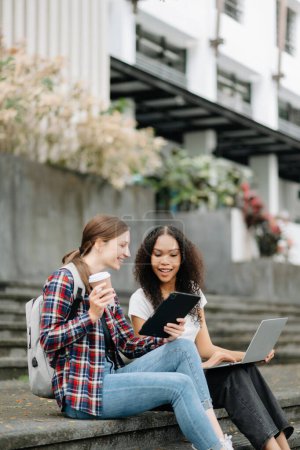 Téléchargez les photos : Jeunes étudiantes se concentrant sur un projet scolaire, regardant un ordinateur portable, buvant du café, discutant et travaillant ensemble au parc du campus - en image libre de droit