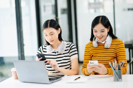 Téléchargez les photos : Deux jeunes filles asiatiques attrayantes qui travaillent sur le projet de l'école en utilisant un ordinateur portable ensemble, profiter de parler le matin - en image libre de droit