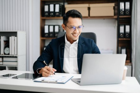 Téléchargez les photos : Jeune asiatique mâle employé de bureau en costume d'entreprise travaillant et souriant dans le bureau - en image libre de droit