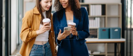 Photo for Happy two young business women holding coffee cups and using smartphone in co-working office - Royalty Free Image