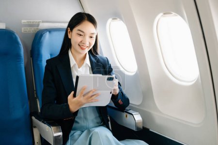 Téléchargez les photos : Attrayant asiatique passager féminin de l'avion assis dans un siège confortable tout en travaillant sur tablette numérique, en utilisant une connexion sans fil. Voyagez avec style, travaillez avec grâce - en image libre de droit