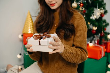 Téléchargez les photos : Joyeux jeune femme tenant boîte cadeau assis près de l'arbre de Noël à la maison - en image libre de droit