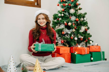 Photo for Happy young woman holding gift box sitting near christmas tree at home - Royalty Free Image