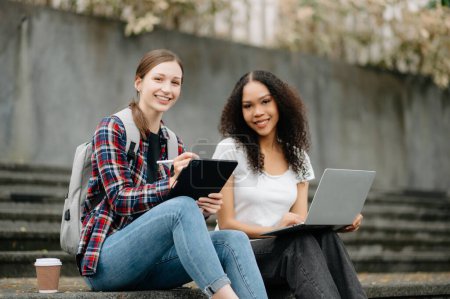 Téléchargez les photos : Jeunes étudiantes se concentrant sur un projet scolaire, regardant un ordinateur portable, buvant du café, discutant et travaillant ensemble au parc du campus - en image libre de droit