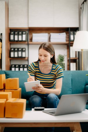 Photo for Young female business owner using laptop, taking notes and preparing parcels for sending online orders to customer in home office. Shopping Online concept - Royalty Free Image