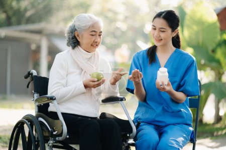 Photo for Young Asian nurse giving pills to senior woman on wheelchair in hospital park. Senior healthcare concept - Royalty Free Image