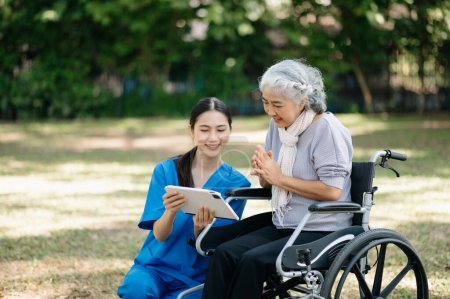 Téléchargez les photos : Personnes âgées asiatiques femme âgée en fauteuil roulant avec soignant asiatique et encourager le patient à utiliser une tablette numérique dans le jardin - en image libre de droit