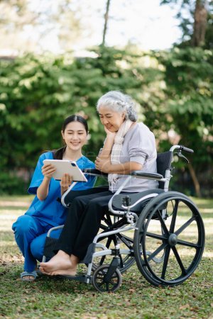 Photo for Elderly asian senior woman on wheelchair with Asian careful caregiver and encourage patient using digital tablet  in garden - Royalty Free Image