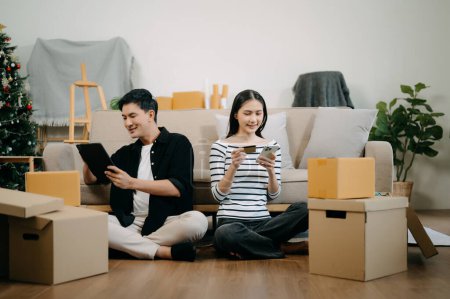 Photo for Asian young attractive couple man and woman using smartphone and digital tablet  for online shopping in new house. Young married asian moving home. - Royalty Free Image