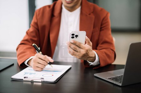 Téléchargez les photos : Homme d'affaires confiant travaillant sur smartphone et écrivant dans des documents sur le lieu de travail au bureau moderne - en image libre de droit