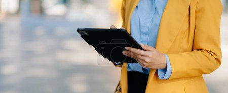 Téléchargez les photos : Jeune femme d'affaires asiatique utilisant une tablette numérique dans la rue dans une grande ville avec un centre d'affaires en arrière-plan - en image libre de droit