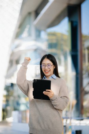 Photo for Young Asian businesswoman using digital tablet on the street in big city with business center on the background - Royalty Free Image