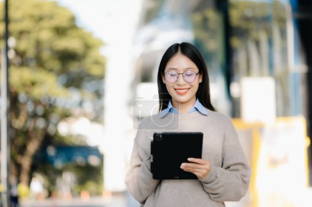 Photo for Confident Asian woman with a smile standing on the street at outside office. Businesswoman using digital tablet with business center on the background - Royalty Free Image