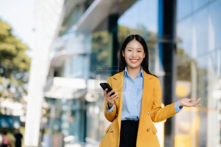 Foto de Joven mujer de negocios asiática usando smartphone en la calle en la gran ciudad con centro de negocios en el fondo - Imagen libre de derechos