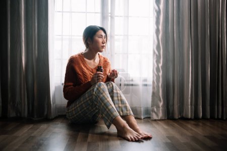 Téléchargez les photos : Femme asiatique se sentant dépression assis sur le sol à la fenêtre avec bouteille de pilules dans la maison - en image libre de droit