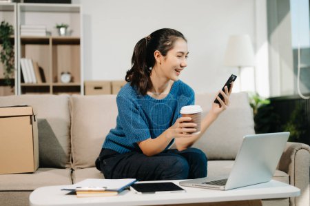 Foto de Mujer de negocios asiática usando un teléfono inteligente con una sonrisa mientras está sentada en el sofá en su oficina en casa - Imagen libre de derechos