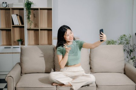 Téléchargez les photos : Jeune femme asiatique à l'aide d'un smartphone assis sur le canapé à la maison - en image libre de droit