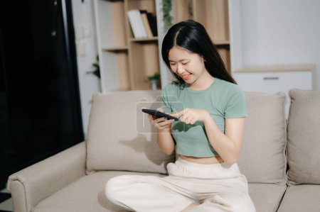 Téléchargez les photos : Jeune femme asiatique à l'aide d'un smartphone assis sur le canapé à la maison - en image libre de droit