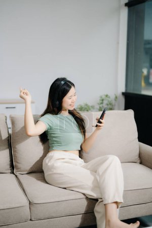 Foto de Joven asiático mujer relajarse en cómodo sofá en casa mensajes de texto en el teléfono inteligente - Imagen libre de derechos