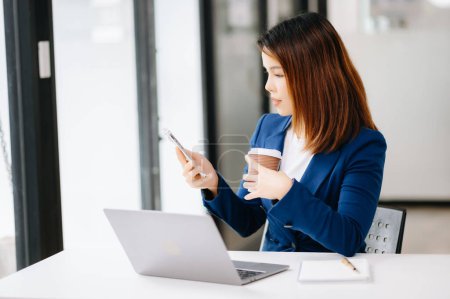 Téléchargez les photos : Jeune femme d'affaires asiatique utilisant un smartphone tout en travaillant dans le bureau - en image libre de droit