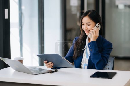 Foto de Mujer de negocios asiática usando teléfono inteligente y portátil con una sonrisa mientras está sentada en la oficina - Imagen libre de derechos