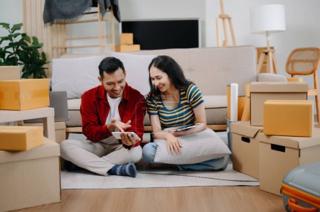 Foto de Feliz asiática joven atractiva pareja hombre y mujer usando smartphone y tableta con grandes cajas de mudanza en una nueva casa - Imagen libre de derechos