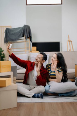 Foto de Feliz asiática joven atractiva pareja hombre y mujer usando smartphone con grandes cajas moviéndose en una nueva casa - Imagen libre de derechos