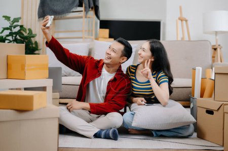 Téléchargez les photos : Heureux asiatique jeune attrayant couple homme et femme en utilisant smartphone avec de grandes boîtes se déplaçant dans une nouvelle maison - en image libre de droit