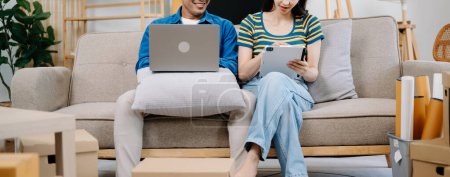 Foto de Asiático joven atractivo pareja hombre y mujer uso portátil y tableta para comprar muebles en línea para decorar nueva casa - Imagen libre de derechos