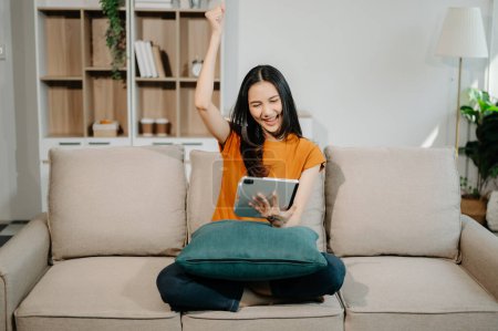Téléchargez les photos : Heureuse femme asiatique en utilisant la tablette numérique sur le canapé à la maison - en image libre de droit
