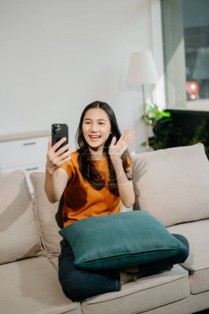 Téléchargez les photos : Heureuse femme asiatique en utilisant le smartphone sur le canapé à la maison - en image libre de droit