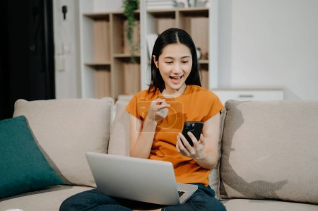 Téléchargez les photos : Jeune femme asiatique se détendre sur un canapé confortable à la maison en utilisant un smartphone et un ordinateur portable - en image libre de droit