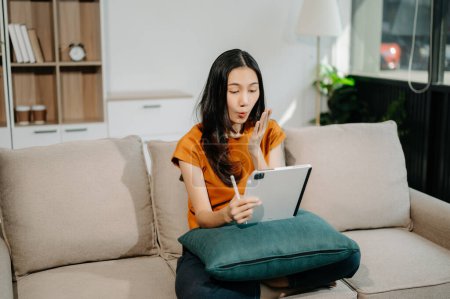 Téléchargez les photos : Jeune femme asiatique se détendre sur un canapé confortable à la maison en utilisant une tablette numérique - en image libre de droit