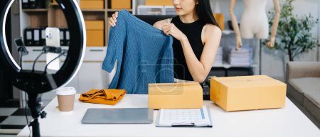 Photo for Fashion blogger concept, Young Asian woman selling clothes on video streaming. Startup small business SME, using smartphone, taking receive and checking in office - Royalty Free Image