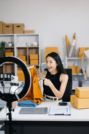 Téléchargez les photos : Concept de blogueur de mode, Jeune femme asiatique vendant des vêtements en streaming vidéo. Démarrage PME de petite entreprise, à l'aide d'un smartphone, réception et enregistrement au bureau - en image libre de droit
