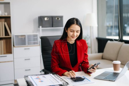 Téléchargez les photos : Asiatique femme d'affaires en utilisant smartphone et ordinateur portable avec un sourire tout en étant assis au bureau - en image libre de droit