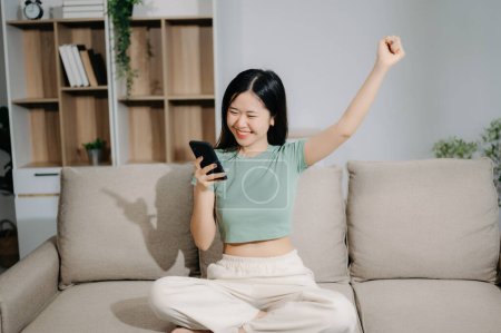 Photo for Happy Young asian woman using smartphone  while seated on couch at home - Royalty Free Image