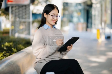 Téléchargez les photos : Jeune femme d'affaires asiatique utilisant un smartphone dans la rue dans une grande ville avec un centre d'affaires en arrière-plan - en image libre de droit