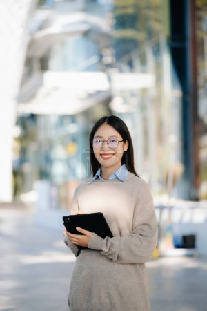 Téléchargez les photos : Femme asiatique confiante avec un sourire debout dans la rue au bureau extérieur. Femme d'affaires utilisant une tablette numérique avec un centre d'affaires en arrière-plan - en image libre de droit
