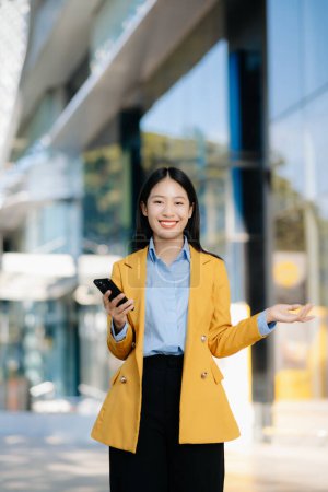 Photo for Young Asian businesswoman using smartphone on the street in big city with business center on the background - Royalty Free Image