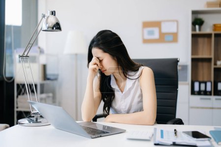 Foto de Mujer asiática sintiendo migraña en la cabeza. Financiera mujer de negocios sobrecargada de trabajo mientras trabajaba en el ordenador portátil en la oficina - Imagen libre de derechos