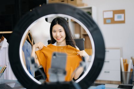 Téléchargez les photos : Belle blogueuse asiatique montrant des vêtements devant la caméra du smartphone pour enregistrer la vidéo vlog en direct en streaming à sa boutique. - en image libre de droit