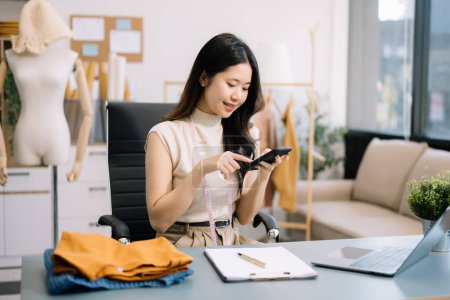 Téléchargez les photos : Femme tailleur asiatique travaillant sur des vêtements dans un atelier de couture. belle jeune créatrice de mode sourire après le succès dans le studio moderne - en image libre de droit